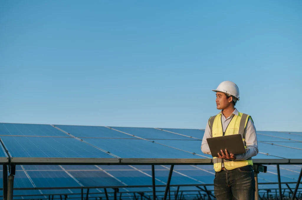 young asian inspector engineer man use laptop computer working solar farm technician supervisor male white helmet checking operation sun photovoltaic solar panel station copy space